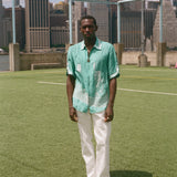 Man wearing the teal stars and stripes cabana with white pants, standing on a soccer field with a city skyline in the background. 