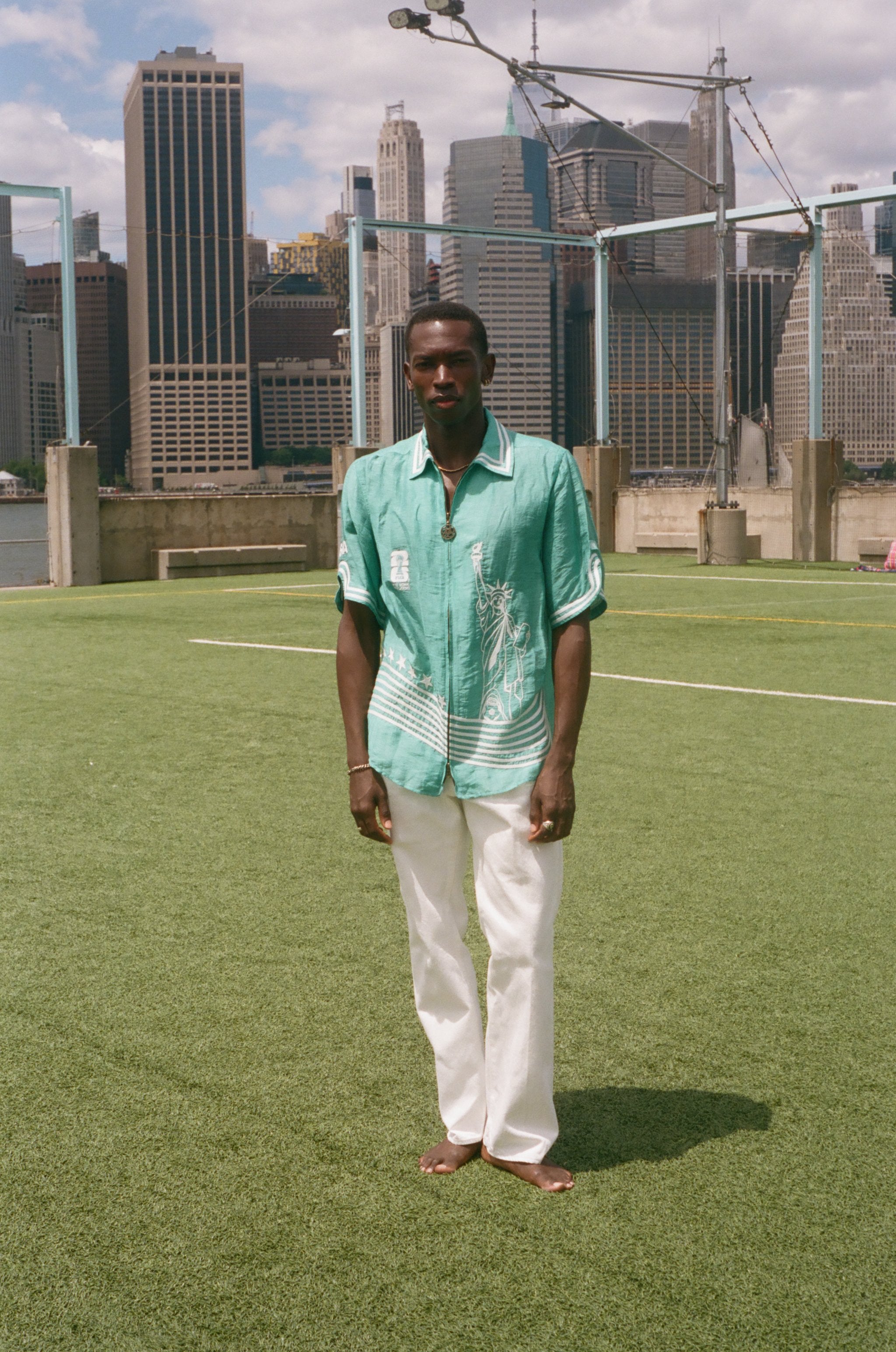 Man wearing the teal stars and stripes cabana with white pants, standing on a soccer field with a city skyline in the background. 