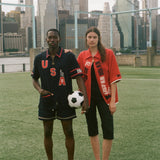 Two people standing on a soccer field with a city skyline in the background, wearing Tombolo and FIFA world cup products. 