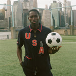 Person holding a soccer ball with a city skyline in the background, wearing the navy USA FIFA cabana shirt. 