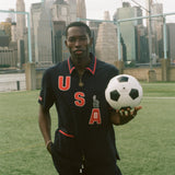 Person holding a soccer ball with a city skyline in the background, wearing the navy USA FIFA cabana shirt. 