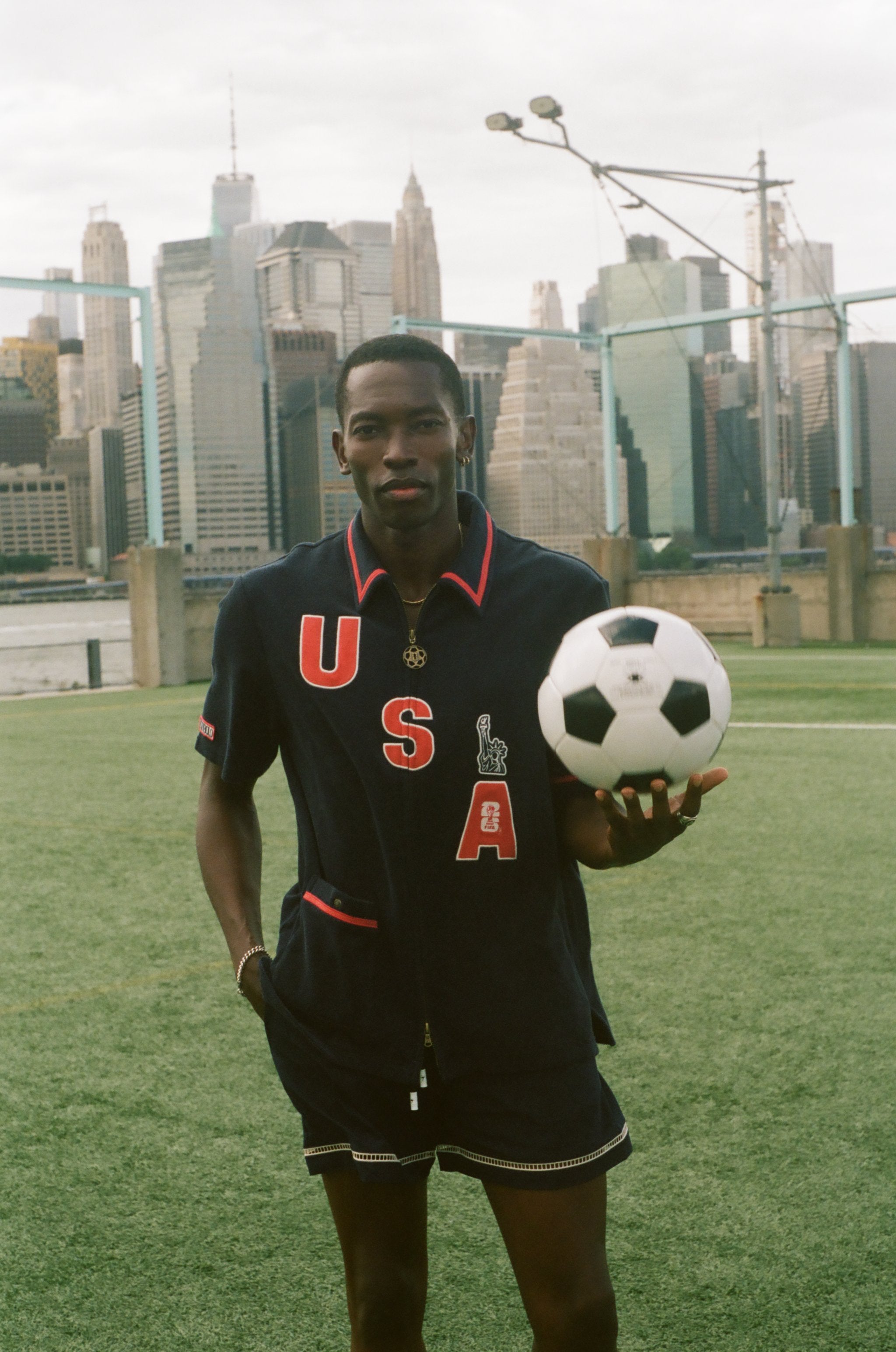 Person holding a soccer ball with a city skyline in the background, wearing the navy USA FIFA cabana shirt. 