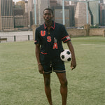 Man holding a soccer ball on a sports field with a city skyline in the background, wearing navy USA FIFA cabana shirt. 