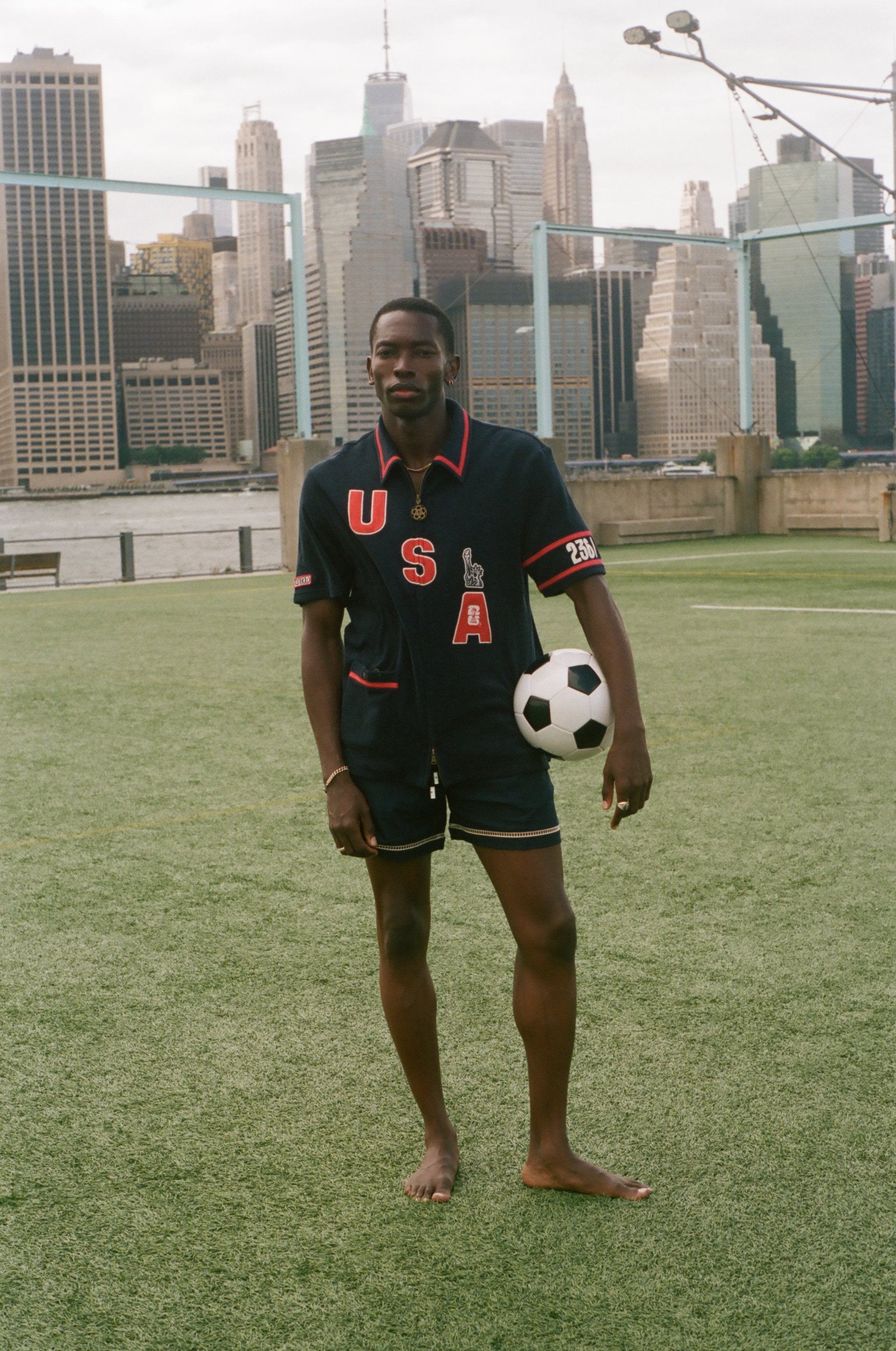 Man holding a soccer ball on a sports field with a city skyline in the background, wearing navy USA FIFA cabana shirt. 