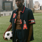 Man holding a soccer ball on a sports field with a city skyline in the background, wearing navy USA FIFA cabana shirt. 