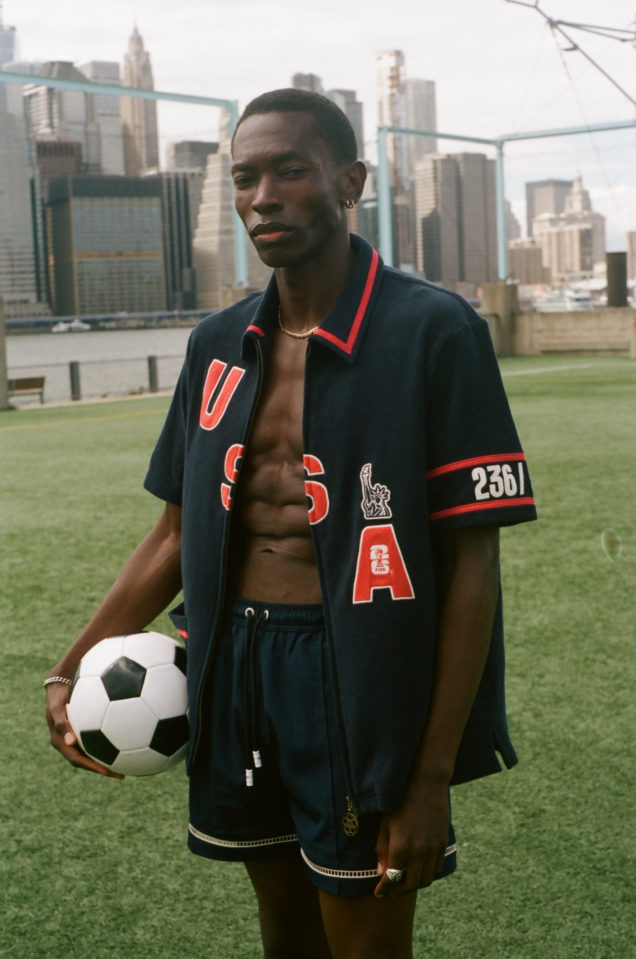 Man holding a soccer ball on a sports field with a city skyline in the background, wearing navy USA FIFA cabana shirt. 