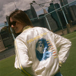 Person wearing a white jacket with graphics and text, standing outdoors with a cityscape in the background.