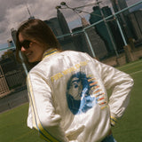 Person wearing a white jacket with graphics and text, standing outdoors with a cityscape in the background.
