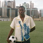Model wearing a cream camp collar shirt with yellow contrast trim, a blue Statue of Liberty graphic, and FIFA World Cup 26 New York New Jersey artwork, standing on a soccer field with the New York City skyline in the background.