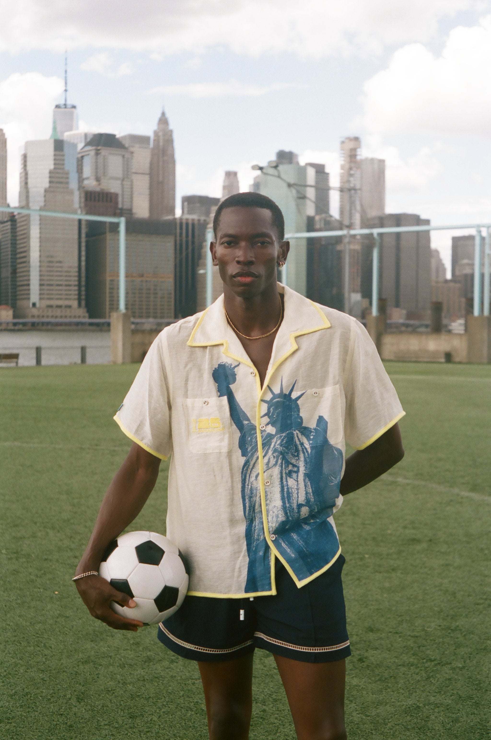 Model wearing a cream camp collar shirt with yellow contrast trim, a blue Statue of Liberty graphic, and FIFA World Cup 26 New York New Jersey artwork, standing on a soccer field with the New York City skyline in the background.