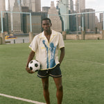 Model wearing a cream camp collar shirt with yellow contrast trim, a blue Statue of Liberty graphic, and FIFA World Cup 26 New York New Jersey artwork, standing on a rsoccer field with the New York City skyline in the background.