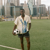 Model wearing a cream camp collar shirt with yellow contrast trim, a blue Statue of Liberty graphic, and FIFA World Cup 26 New York New Jersey artwork, standing on a rsoccer field with the New York City skyline in the background.