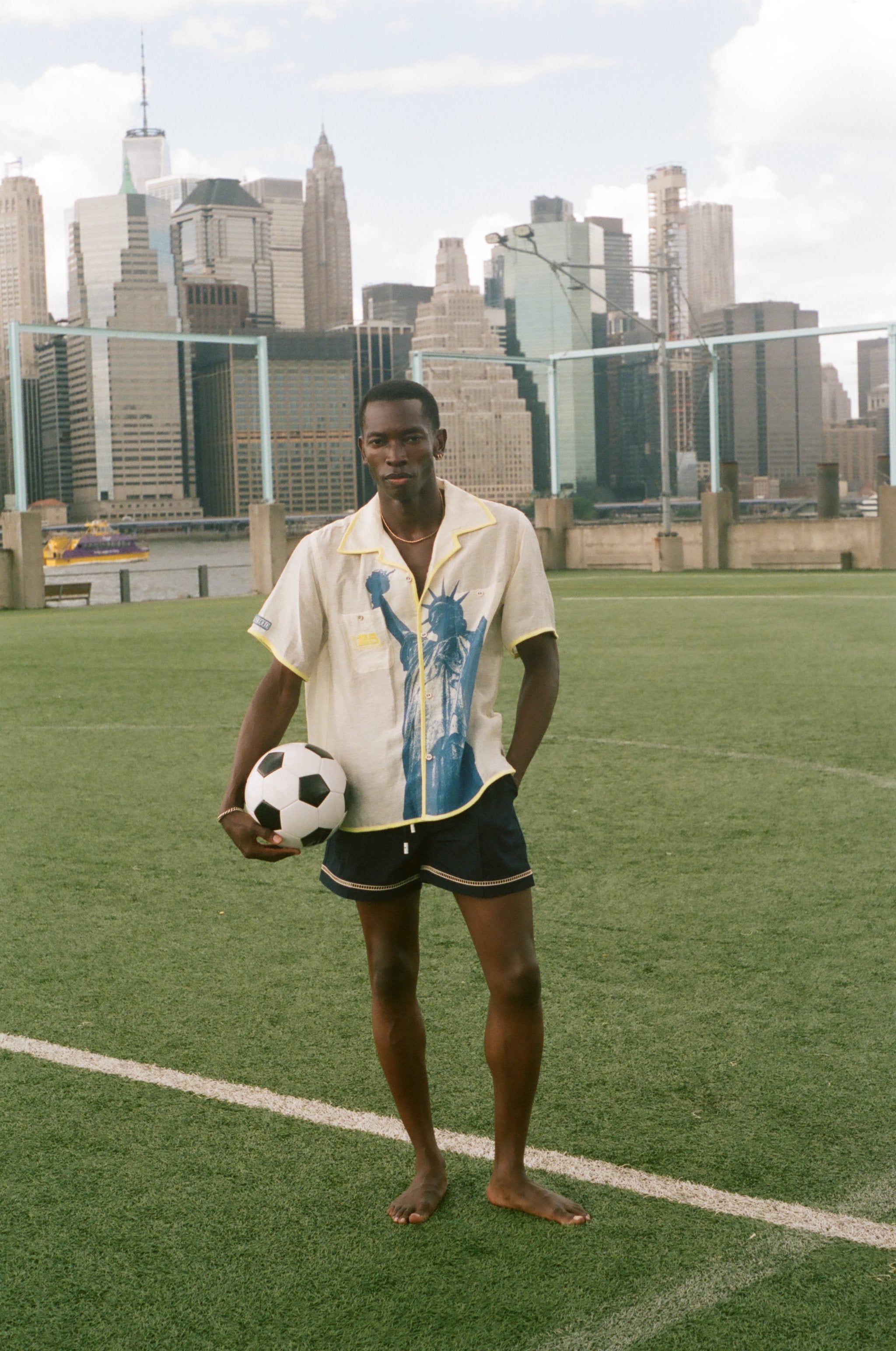 Model wearing a cream camp collar shirt with yellow contrast trim, a blue Statue of Liberty graphic, and FIFA World Cup 26 New York New Jersey artwork, standing on a rsoccer field with the New York City skyline in the background.