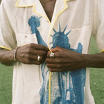 Close up of male model wearing a cream camp collar shirt with yellow contrast trim, a blue Statue of Liberty graphic, and FIFA World Cup 26 New York New Jersey artwork, standing on a rooftop field with the New York City skyline in the background.