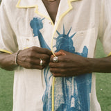 Close up of male model wearing a cream camp collar shirt with yellow contrast trim, a blue Statue of Liberty graphic, and FIFA World Cup 26 New York New Jersey artwork, standing on a rooftop field with the New York City skyline in the background.