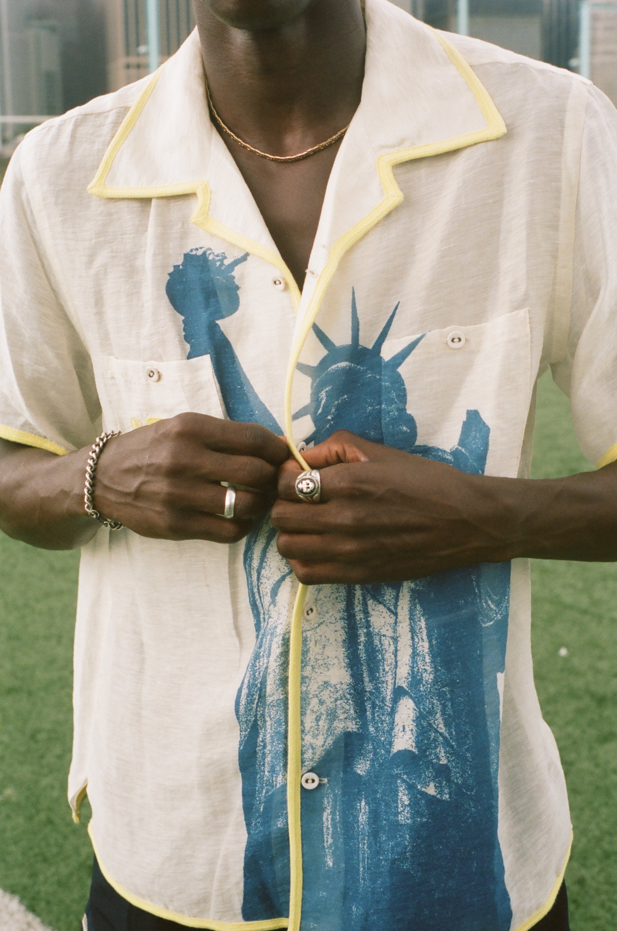 Close up of male model wearing a cream camp collar shirt with yellow contrast trim, a blue Statue of Liberty graphic, and FIFA World Cup 26 New York New Jersey artwork, standing on a rooftop field with the New York City skyline in the background.