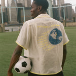 Model holding a soccer ball looking off camera while wearing the beige camp collar FIFA Lady Liberty shirt. 