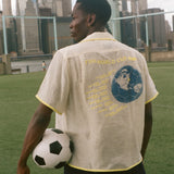 Model holding a soccer ball looking off camera while wearing the beige camp collar FIFA Lady Liberty shirt. 