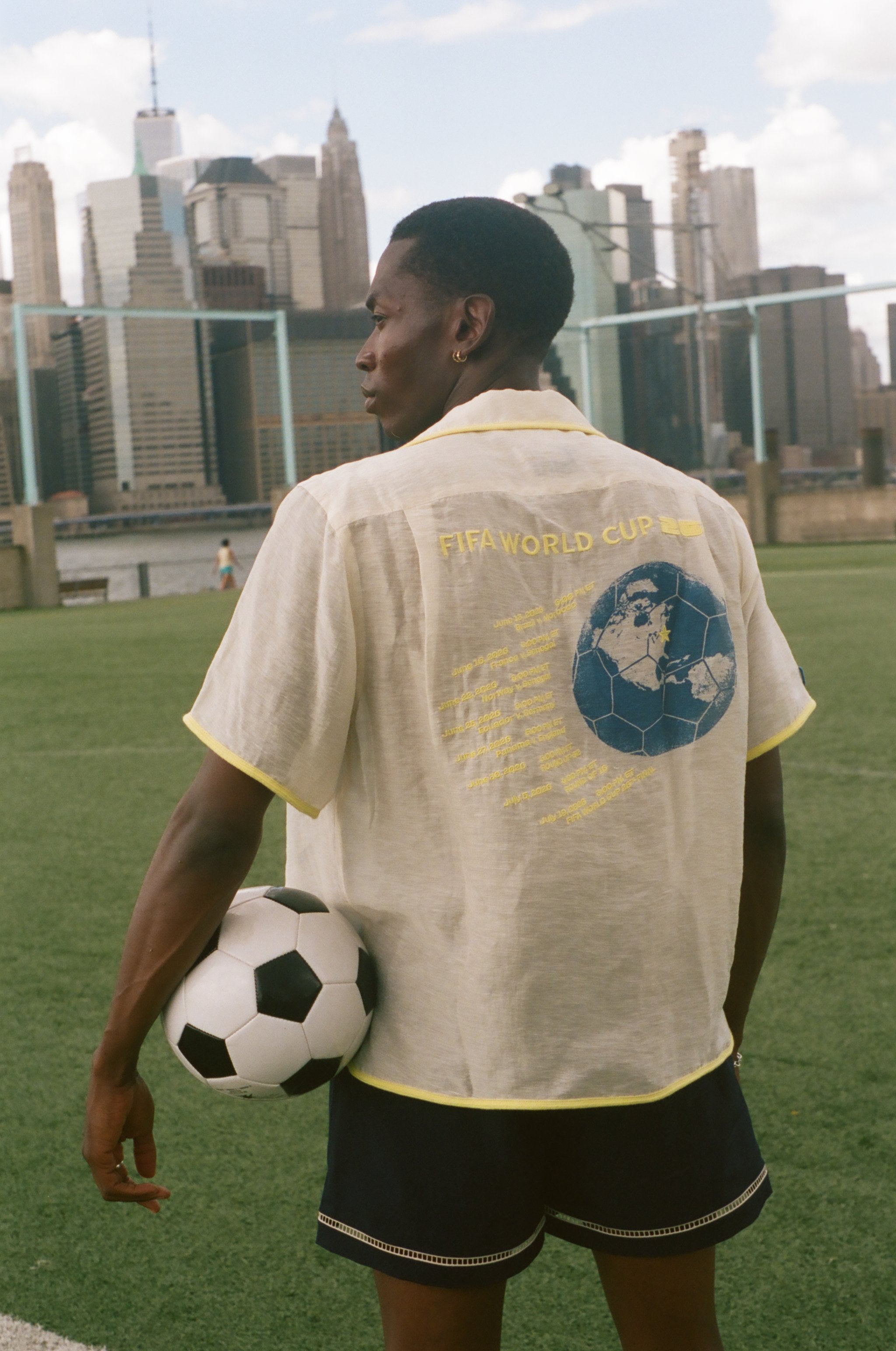 Model holding a soccer ball looking off camera while wearing the beige camp collar FIFA Lady Liberty shirt. 