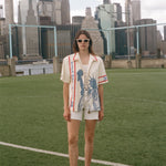 Model wearing a cream terry cloth zip-front shirt with red trim and a blue Statue of Liberty graphic, standing on a soccer field with a soccer ball and the New York City skyline in the background.