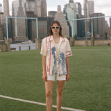 Model wearing a cream terry cloth zip-front shirt with red trim and a blue Statue of Liberty graphic, standing on a soccer field with a soccer ball and the New York City skyline in the background.