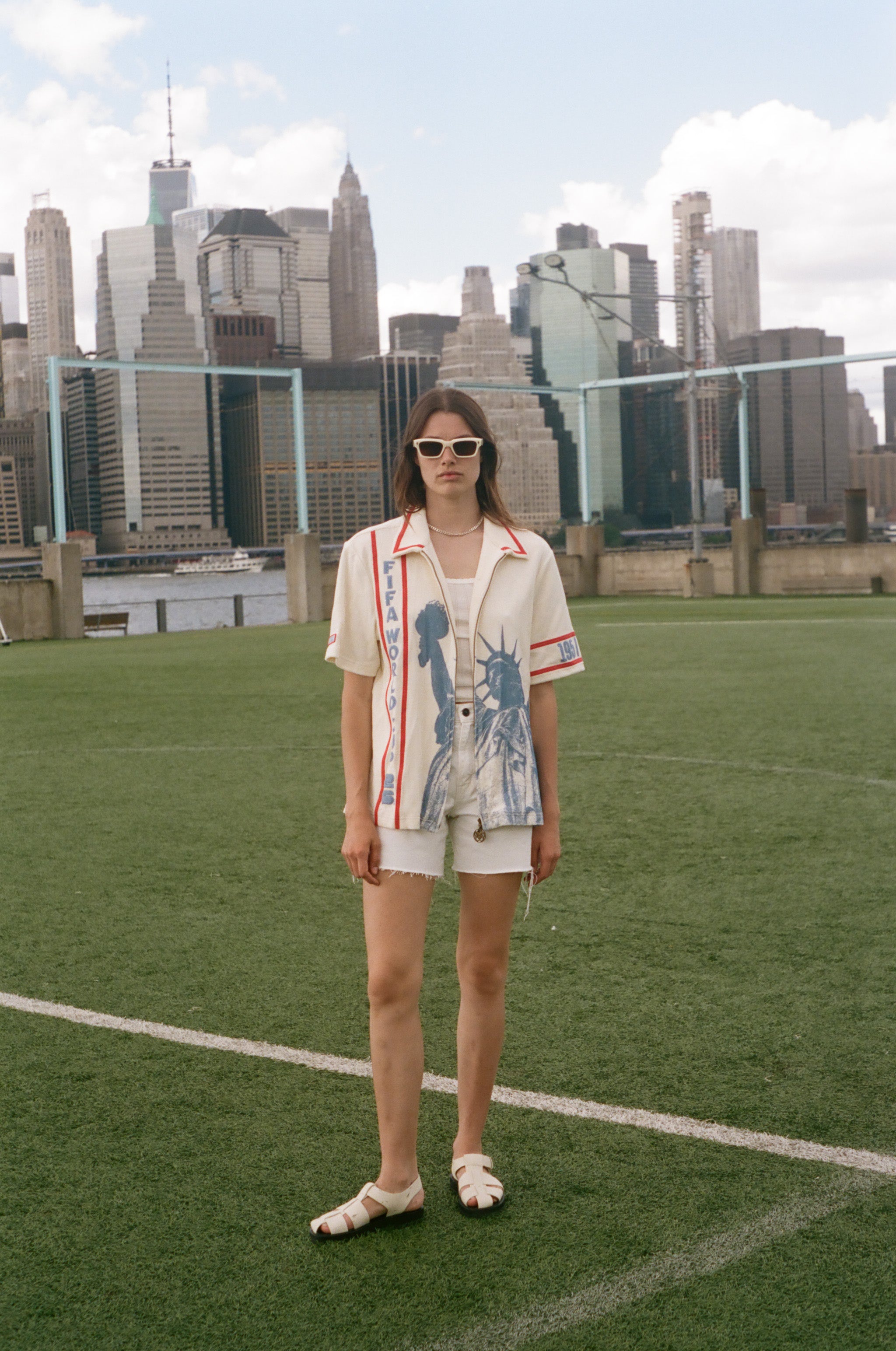 Model wearing a cream terry cloth zip-front shirt with red trim and a blue Statue of Liberty graphic, standing on a soccer field with a soccer ball and the New York City skyline in the background.