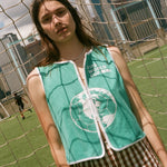 Woman wearing teal FIFA hand towel top, leaning against a soccer goal net. 