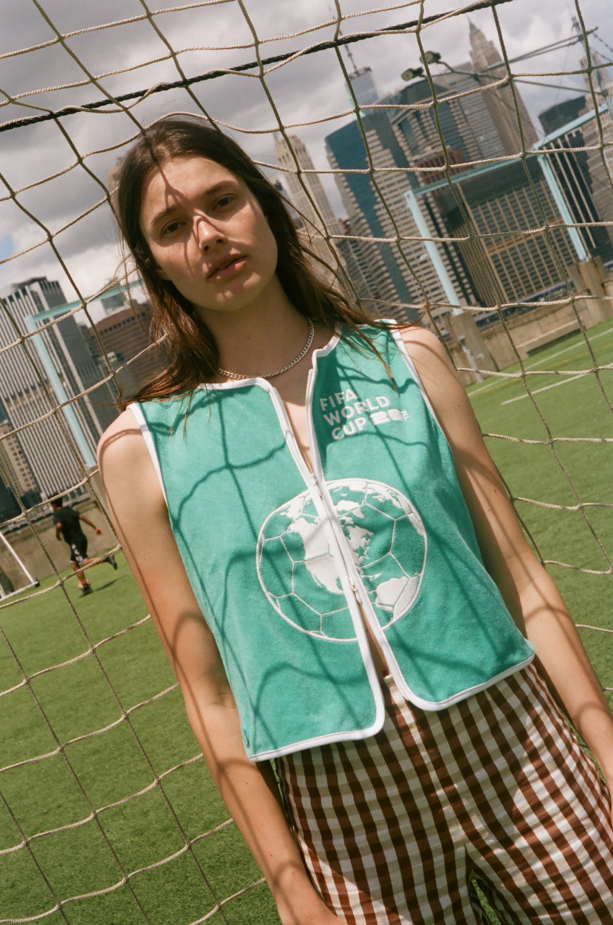 Woman wearing teal FIFA hand towel top, leaning against a soccer goal net. 
