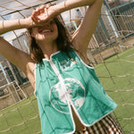 Woman wearing teal FIFA hand towel top, leaning against a soccer goal net. 