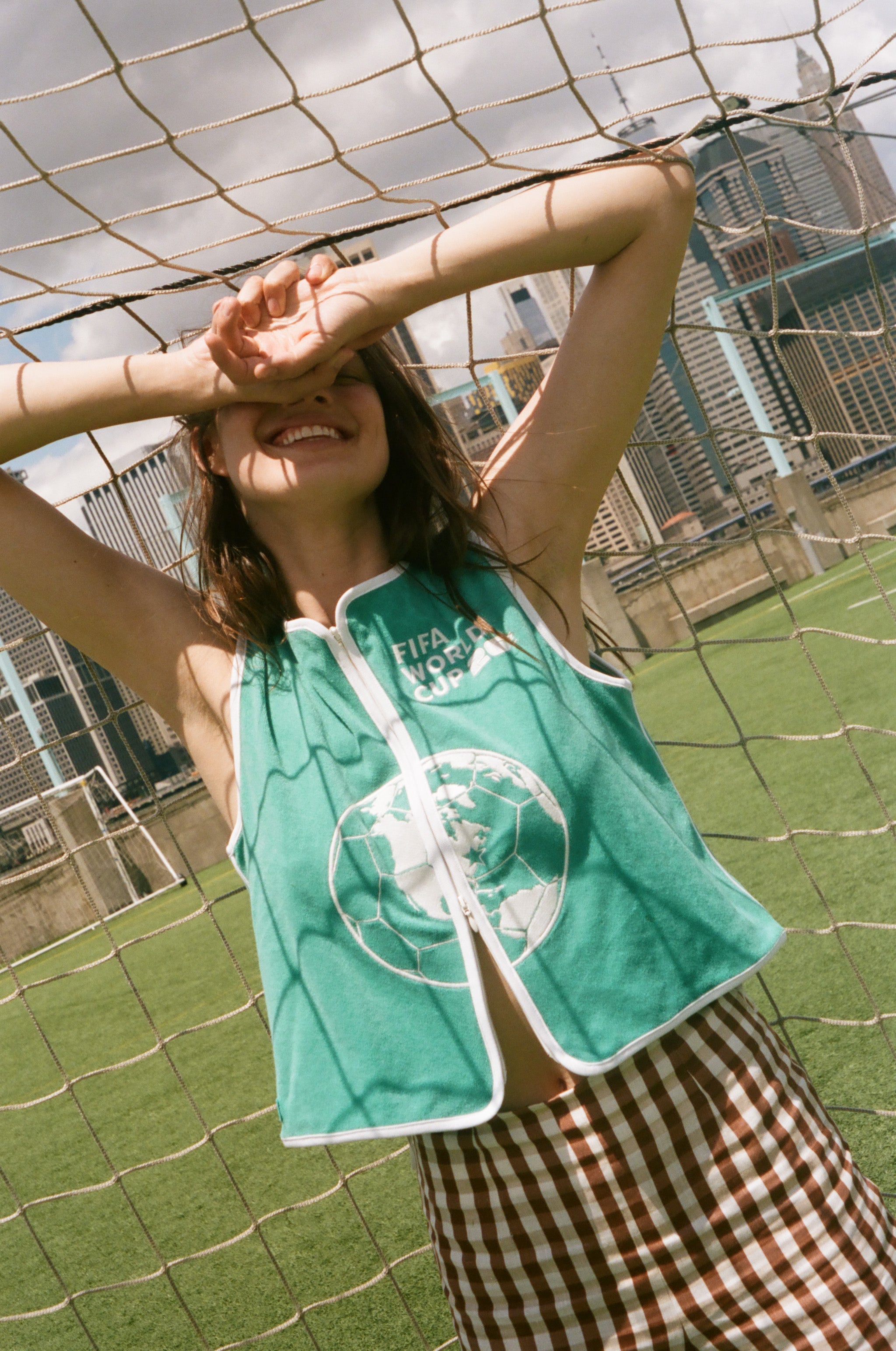Woman wearing teal FIFA hand towel top, leaning against a soccer goal net. 