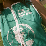 Close-up of  woman wearing teal FIFA hand towel top, leaning against a soccer goal net. 