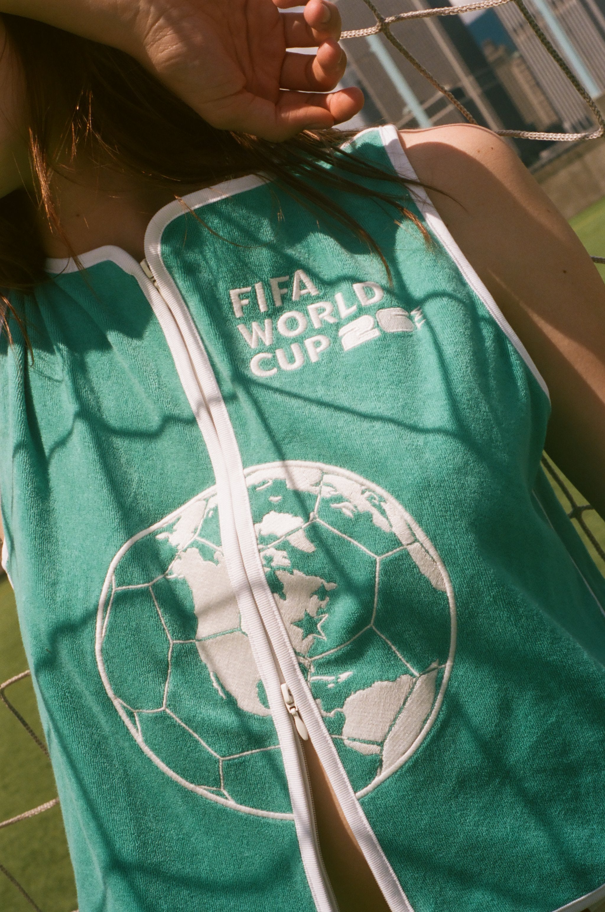 Close-up of  woman wearing teal FIFA hand towel top, leaning against a soccer goal net. 