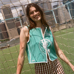 Woman wearing teal FIFA hand towel top, leaning against a soccer goal net. 