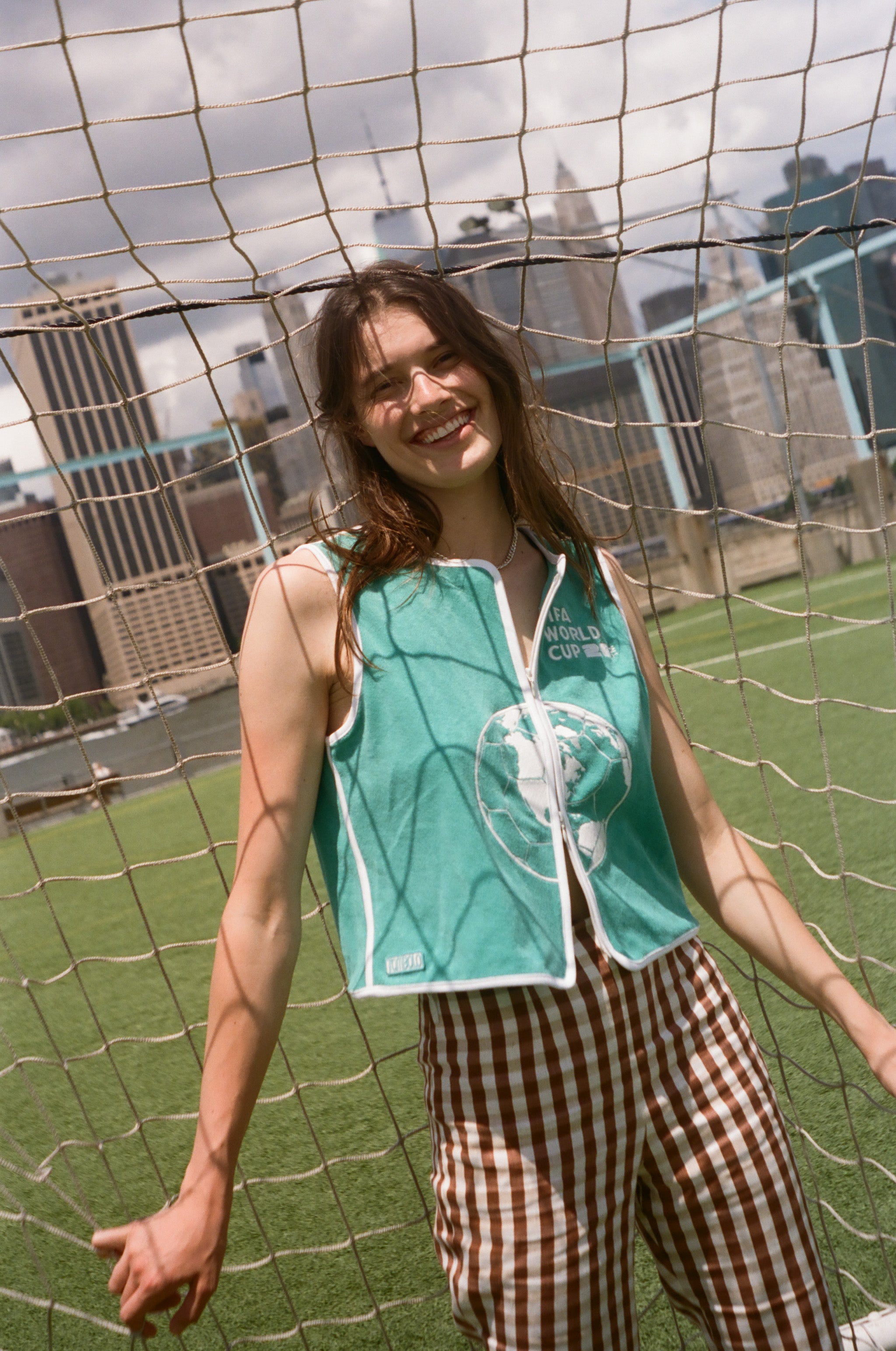 Woman wearing teal FIFA hand towel top, leaning against a soccer goal net. 