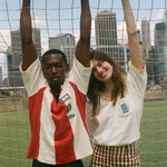 Two people posing in front of a soccer goal with city skyline in the background, wearing items from the FIFA and Tombolo collection.