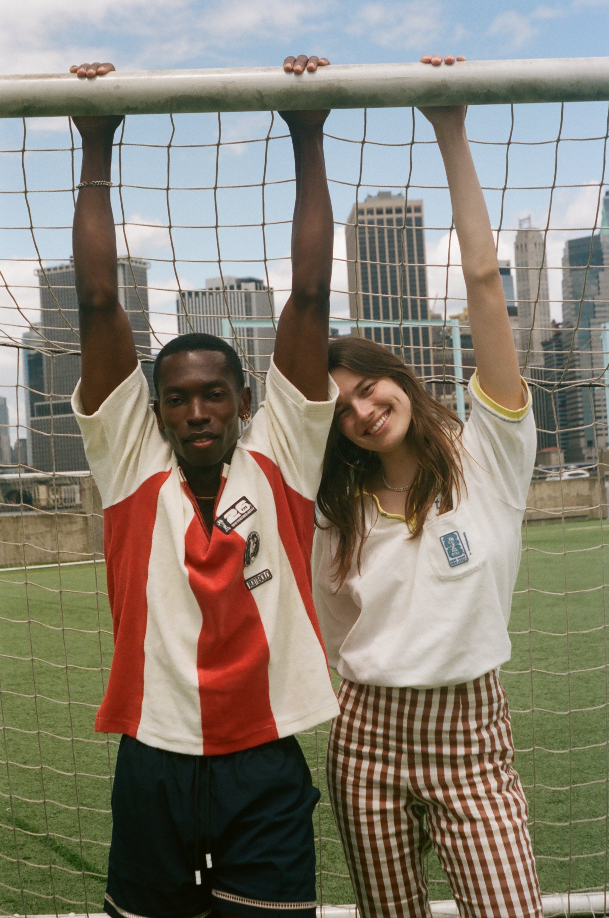 Two people posing in front of a soccer goal with city skyline in the background, wearing items from the FIFA and Tombolo collection.