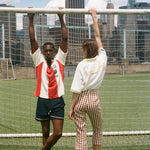 Two people posing in front of a soccer goal with city skyline in the background, wearing items from the FIFA and Tombolo collection.