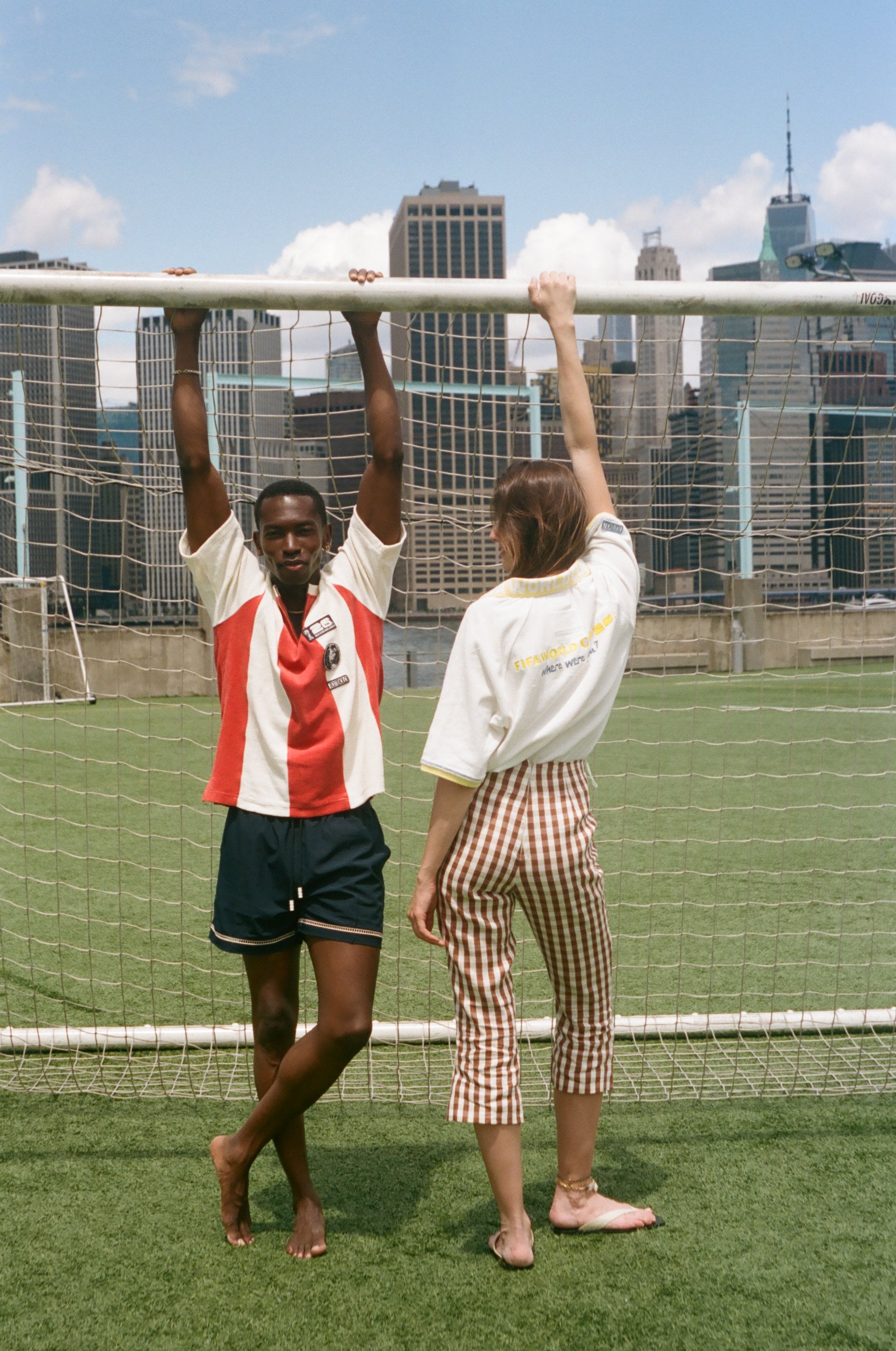 Two people posing in front of a soccer goal with city skyline in the background, wearing items from the FIFA and Tombolo collection.