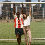 Two people posing in front of a soccer goal with city skyline in the background, wearing items from the FIFA and Tombolo collection.