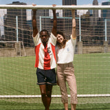 Two people posing in front of a soccer goal with city skyline in the background, wearing items from the FIFA and Tombolo collection.
