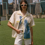 Model wearing a cream camp collar shirt with yellow contrast trim, a blue Statue of Liberty graphic, and FIFA World Cup 26 New York New Jersey artwork, standing on a soccer field with the New York City skyline in the background.