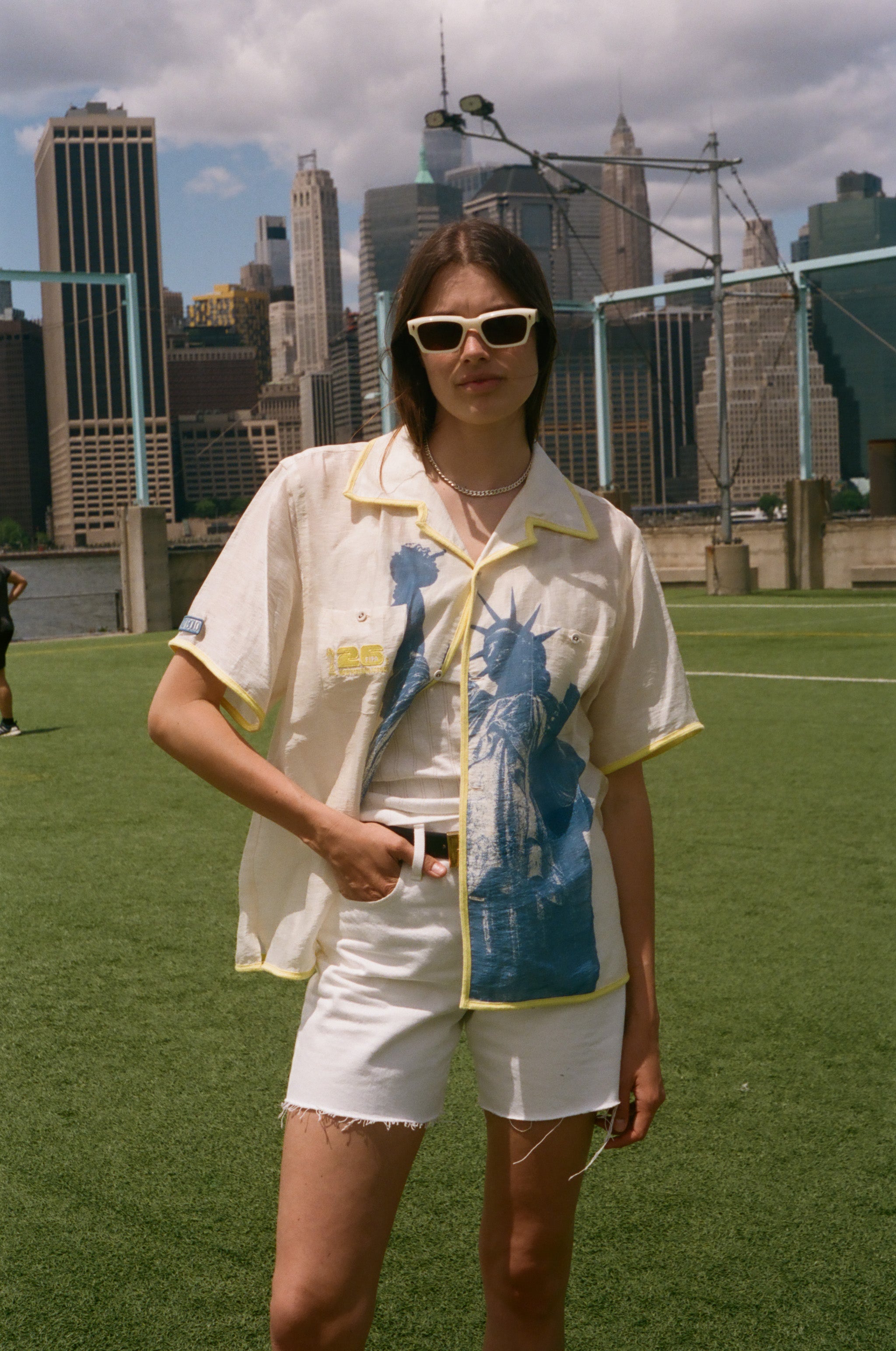 Model wearing a cream camp collar shirt with yellow contrast trim, a blue Statue of Liberty graphic, and FIFA World Cup 26 New York New Jersey artwork, standing on a soccer field with the New York City skyline in the background.