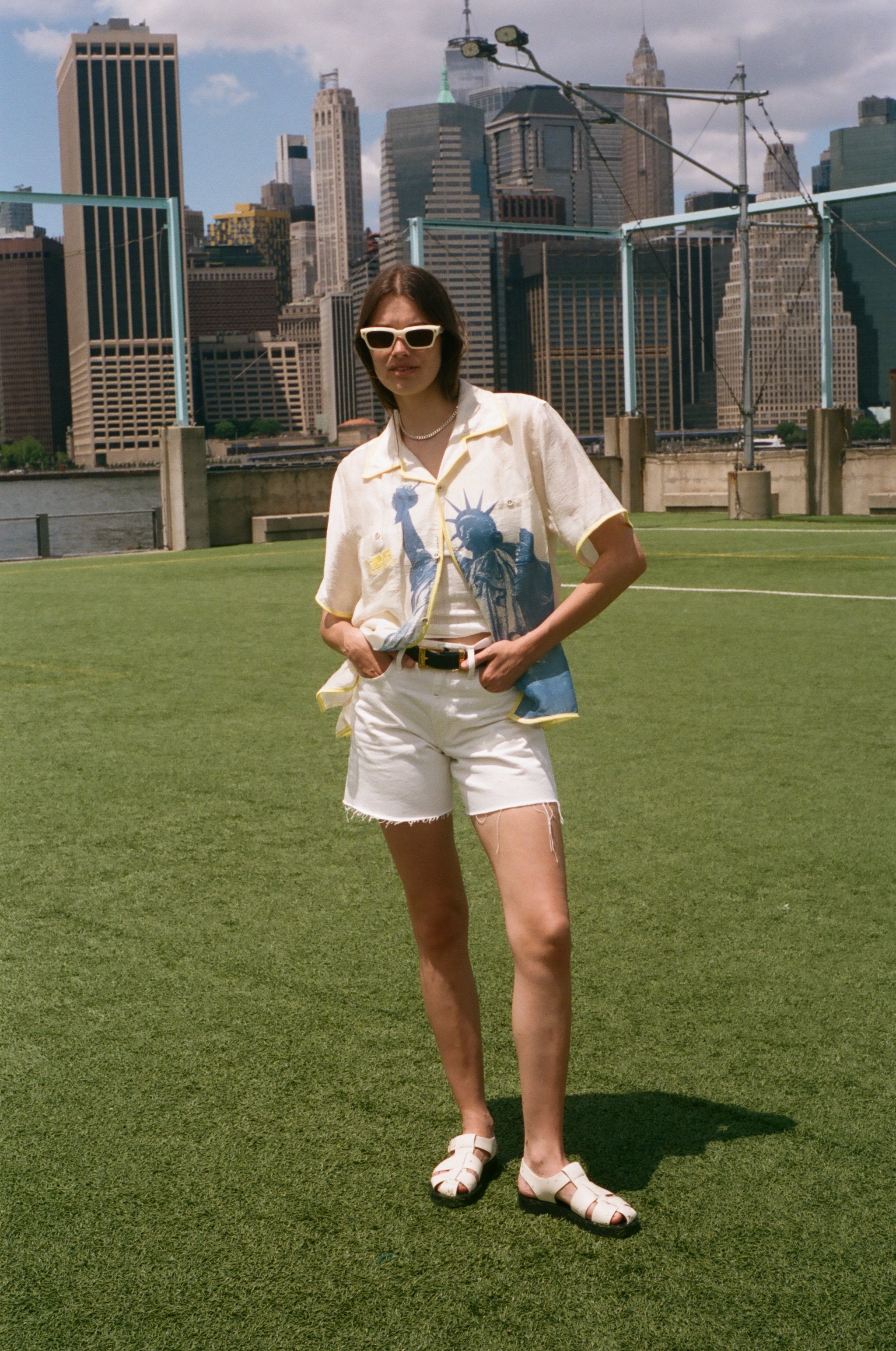 Woman in a white outfit with a blue statue of liberty design on her shirt for FIFA World Cup 2026 standing on grass with a city skyline in the background
