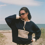 A woman posing on a sandy beach wearing a navy blue long-sleeve knit sweater with cream trim and a front pocket, paired with black and white striped shorts.