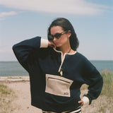 A woman posing on a sandy beach wearing a navy blue long-sleeve knit sweater with cream trim and a front pocket, paired with black and white striped shorts.