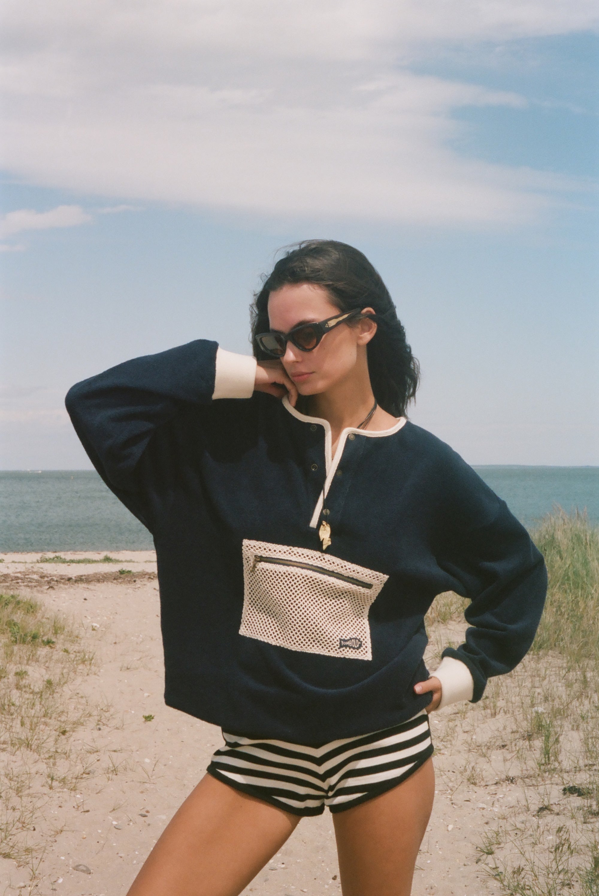 A woman posing on a sandy beach wearing a navy blue long-sleeve knit sweater with cream trim and a front pocket, paired with black and white striped shorts.