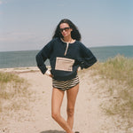 A woman stands on a sandy beach path wearing a navy blue sweatshirt with a beige pocket detail, black and white striped shorts, and black sandals, with the ocean and sky in the background.