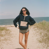 A woman stands on a sandy beach path wearing a navy blue sweatshirt with a beige pocket detail, black and white striped shorts, and black sandals, with the ocean and sky in the background.