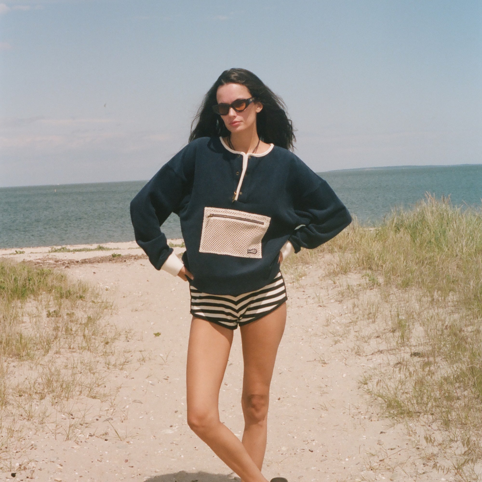 A woman stands on a sandy beach path wearing a navy blue sweatshirt with a beige pocket detail, black and white striped shorts, and black sandals, with the ocean and sky in the background.