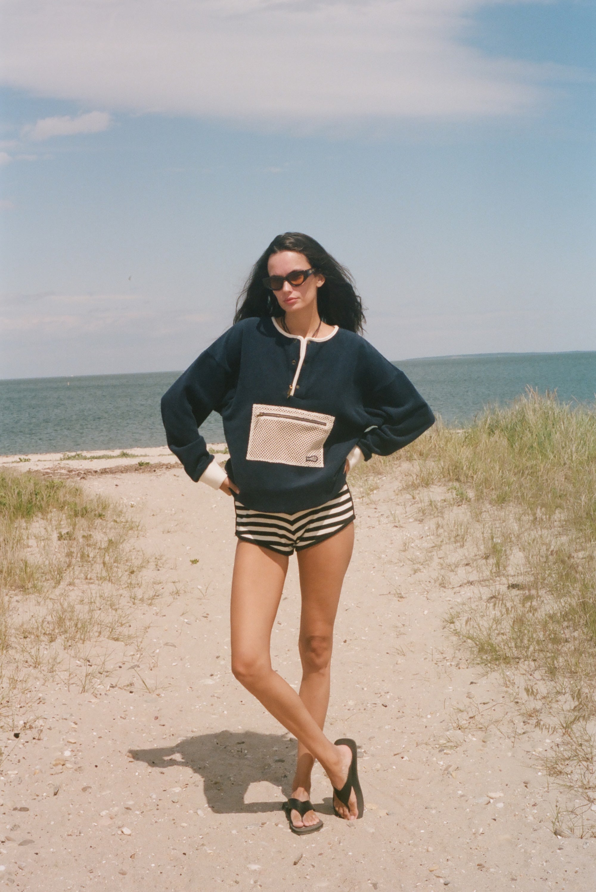 A woman stands on a sandy beach path wearing a navy blue sweatshirt with a beige pocket detail, black and white striped shorts, and black sandals, with the ocean and sky in the background.